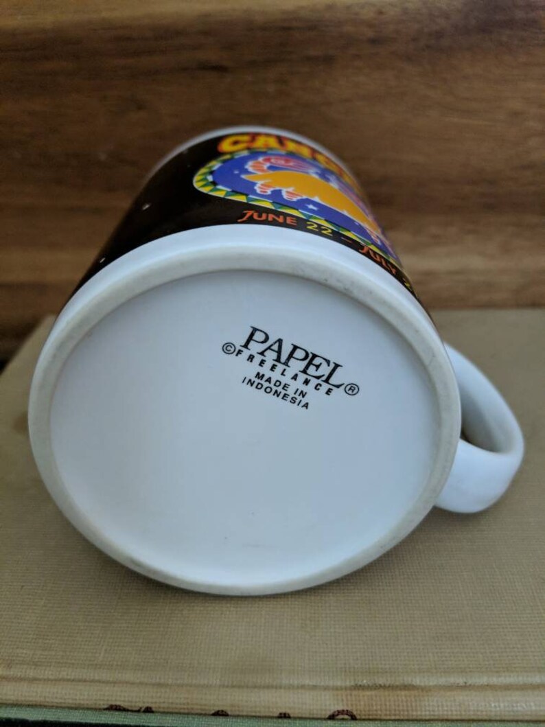 May include: White ceramic mug with a black upper band featuring the word "CAMEL" and the dates "JUNE 22 - JULY". The bottom of the mug has the text "PAPEL FREELANCE MADE IN INDONESIA".