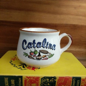 May include: A white ceramic mug with the name "Catalina" in blue script, decorated with colorful vegetable illustrations. The mug has a brown rim and handle, resting on a yellow book.