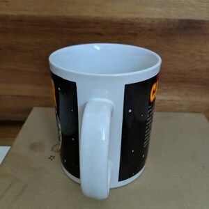 May include: White ceramic coffee mug with a black background featuring a colorful graphic and text. The mug has a white handle and interior. The design includes stars and a yellow and orange graphic.