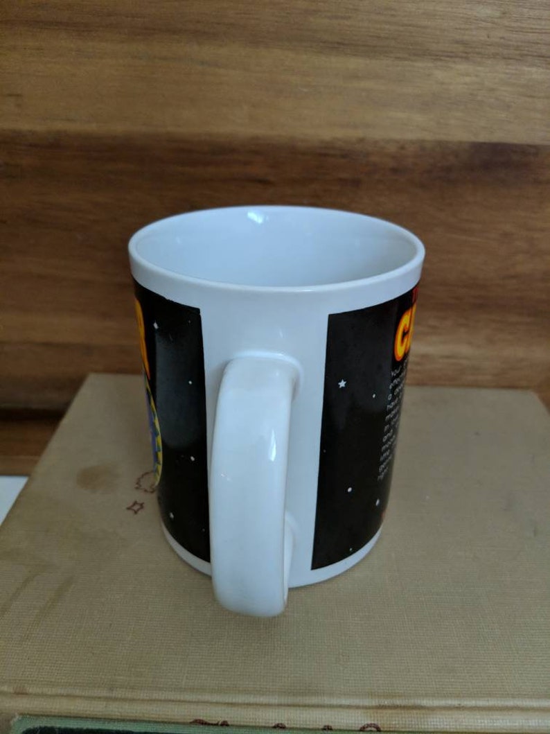 May include: White ceramic coffee mug with a black background featuring a colorful graphic and text. The mug has a white handle and interior. The design includes stars and a yellow and orange graphic.