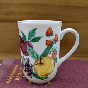 May include: White ceramic mug with a gold rim, adorned with a colorful fruit design. The illustration includes plums, grapes, strawberries, and an apple. The mug has a curved handle and is set on a book with the word "BRIGHT" visible.