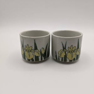 May include: Two ceramic cups with a floral design. The cups are cylindrical with a light gray base and a yellow iris flower design with green leaves. The cups are displayed on a white surface.