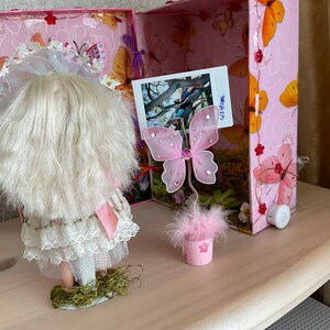 May include: A pink and white doll with blonde hair and a white lace dress is standing on a green base. A pink butterfly with glitter accents is on a pink feathered base in a pink pot. The doll and butterfly are in front of a pink and white box with a photo of a tree and people.