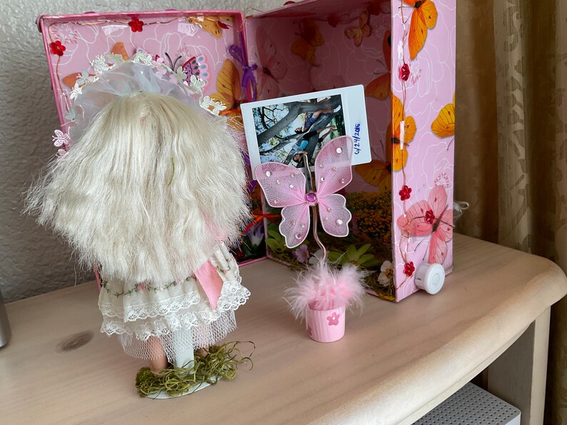 May include: A pink and white doll with blonde hair and a white lace dress is standing on a green base. A pink butterfly with glitter accents is on a pink feathered base in a pink pot. The doll and butterfly are in front of a pink and white box with a photo of a tree and people.