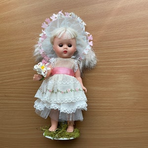 May include: A vintage porcelain doll wearing a white dress with pink accents and a pink and white flower hat. The doll is holding a white flower and is standing on a green base.