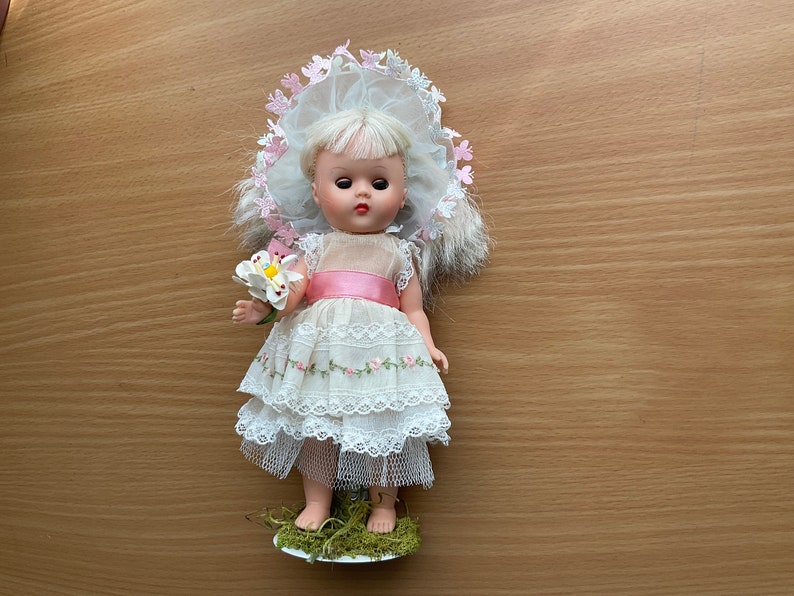 May include: A vintage porcelain doll wearing a white dress with pink accents and a pink and white flower hat. The doll is holding a white flower and is standing on a green base.