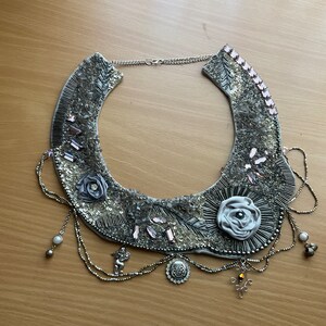May include: A silver and gray statement necklace with a large, white fabric rose and smaller fabric roses. The necklace has a chain with multiple strands and a silver pendant.