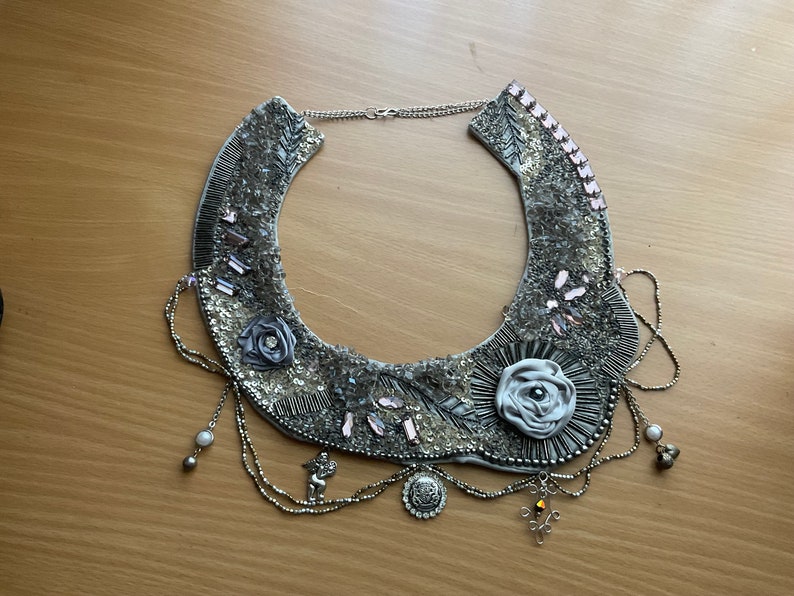 May include: A silver and gray statement necklace with a large, white fabric rose and smaller fabric roses. The necklace has a chain with multiple strands and a silver pendant.