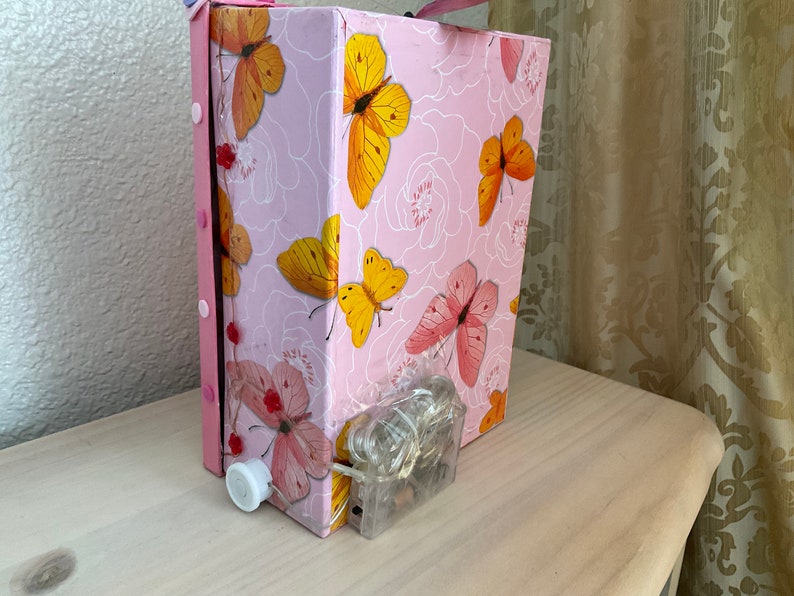May include: A pink cardboard box with a butterfly pattern. The box has a white switch on the side and a clear plastic cord with lights coming out of the bottom.