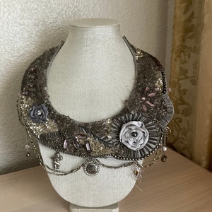 May include: A silver and gray beaded necklace with a large flower design. The necklace is on a white mannequin bust.
