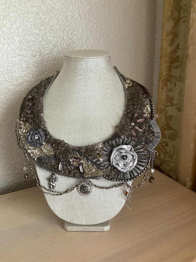 May include: A silver and gray beaded necklace with a large flower design. The necklace is on a white mannequin bust.