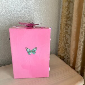 May include: A pink wooden box with a butterfly design. The box has a light blue butterfly on the front and a pink butterfly on the top.