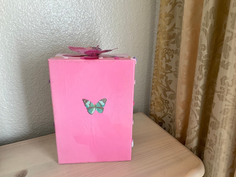May include: A pink wooden box with a butterfly design. The box has a light blue butterfly on the front and a pink butterfly on the top.