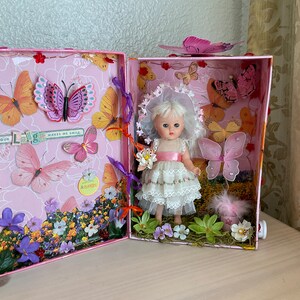 May include: A pink and white dollhouse with a miniature doll wearing a white dress and a pink sash. The dollhouse is decorated with butterflies, flowers, and the words "Your Love Makes Me Smile".