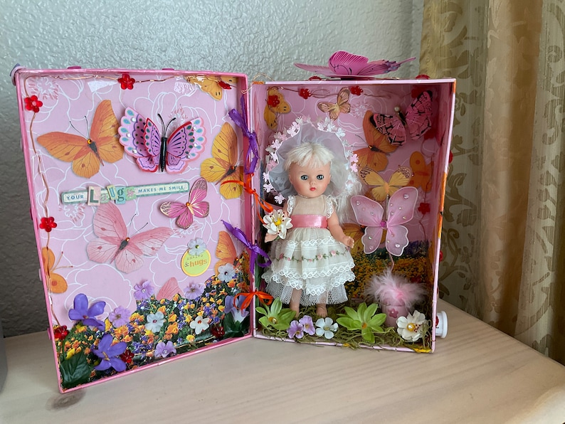 May include: A pink and white dollhouse with a miniature doll wearing a white dress and a pink sash. The dollhouse is decorated with butterflies, flowers, and the words "Your Love Makes Me Smile".