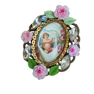 May include: A gold-toned oval brooch with a cameo of a cherub holding an arrow. The cameo is surrounded by pink roses, green leaves, and clear rhinestones.