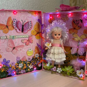 May include: A pink and white decorated box with a miniature doll wearing a white dress and a pink sash. The box is decorated with butterflies, flowers, and lights. The text "Your Laugh Makes Me Smile" is visible on the box.