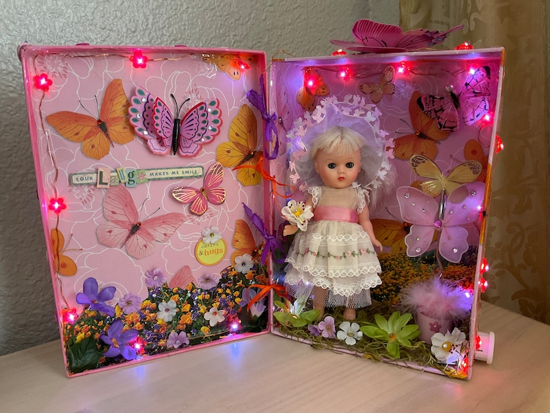 May include: A pink and white decorated box with a miniature doll wearing a white dress and a pink sash. The box is decorated with butterflies, flowers, and lights. The text "Your Laugh Makes Me Smile" is visible on the box.