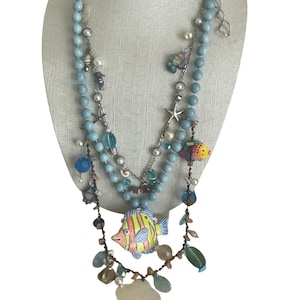 May include: A multi-strand necklace with blue beads, silver charms, and colorful fish pendants. The necklace features a large, yellow and blue fish pendant with a white flower charm hanging below.