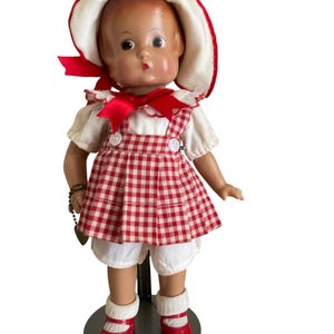 May include: A vintage-style doll with brown hair, wearing a white bonnet with red trim, a red and white gingham pinafore dress over a white short-sleeved shirt, white bloomers, white socks, and shiny red shoes, featuring a small metal heart charm.