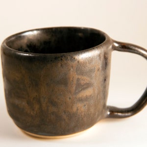 May include: A dark brown ceramic mug with a slightly rounded shape and a handle. The mug has a speckled glaze with a metallic sheen.