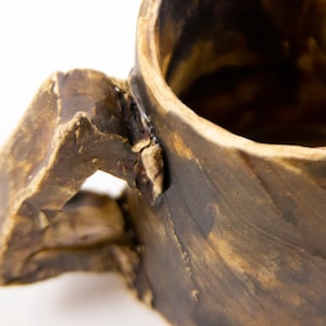 Könnte beinhalten: Nahaufnahme einer handgefertigten Keramiktasse mit strukturierter, rauer Oberfläche. Die Tasse ist überwiegend braun mit dunkleren braunen Akzenten und einem stabilen Henkel. Das Innere ist sichtbar, was darauf hindeutet, dass sie gebrauchsfertig ist.