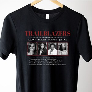 May include: Black t-shirt featuring the word "TRAILBLAZERS" in red, with the words "LEGACY LEADER ACTIVIST JUSTICE" below. The shirt includes images of Michelle Obama, Kamala Harris, Stacey Abrams, and Ketanji Brown Jackson, with inspirational quotes.