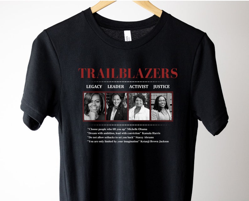 May include: Black t-shirt featuring the word "TRAILBLAZERS" in red, with the words "LEGACY LEADER ACTIVIST JUSTICE" below. The shirt includes images of Michelle Obama, Kamala Harris, Stacey Abrams, and Ketanji Brown Jackson, with inspirational quotes.