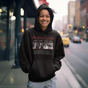 May include: Black hooded sweatshirt with the word "TRAILBLAZERS" in red letters. Below the word are four images of women with the words "LEGACY, LEAD, ACTION, JUSTICE". The sweatshirt is worn by a person standing on a sidewalk.