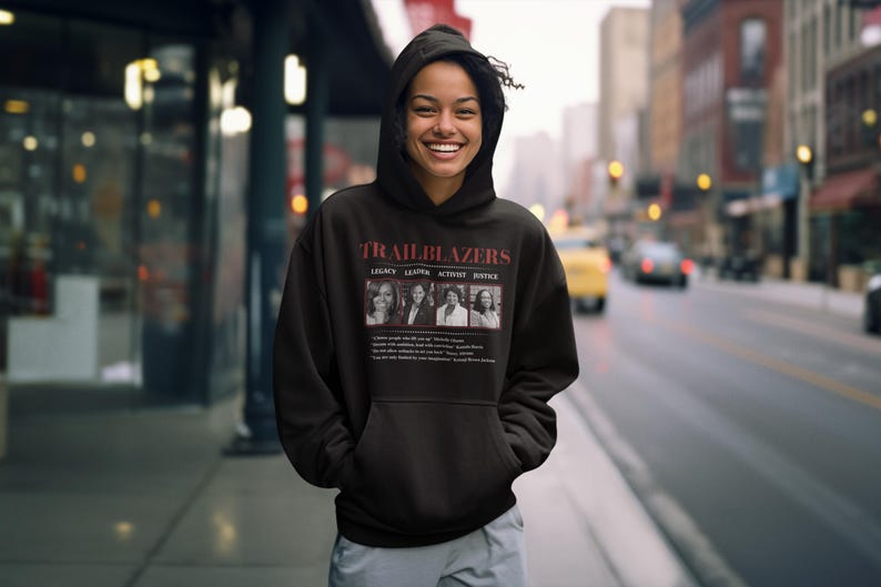 May include: Black hooded sweatshirt with the word "TRAILBLAZERS" in red letters. Below the word are four images of women with the words "LEGACY, LEAD, ACTION, JUSTICE". The sweatshirt is worn by a person standing on a sidewalk.