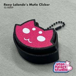 May include: A black and hot pink cat-shaped clicker with a textured surface, featuring white eyes and a mouth. The text Roxy Lalonde's Mutie Clicker CL-0009 is visible at the top, and a black ball chain is attached to the side of the device.