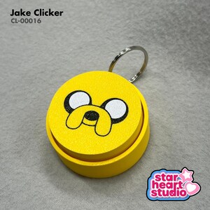 May include: A yellow Jake Clicker keychain with a cartoon face design. The keychain has a silver ring and the text "Jake Clicker" and "CL-00016" is visible. The Star Heart Studio logo is in the bottom right corner.