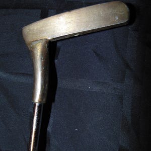 以下が含まれることがあります： 真鍮製のヘッドと銀色のシャフトが付いた、ヴィンテージのゴルフパター。 パターヘッドは、丸みを帯びた端を持つ長方形の形状をしています。 シャフトは、わずかな角度でヘッドに取り付けられています。 パターは暗い背景に配置されています。