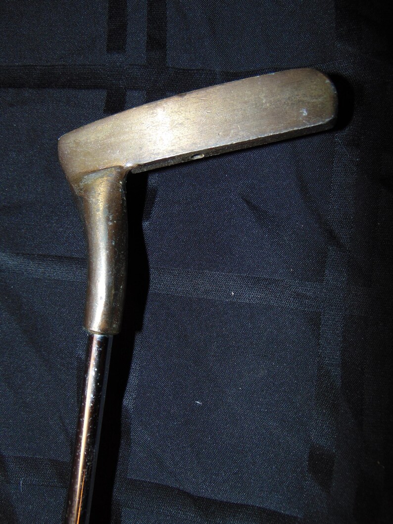 以下が含まれることがあります： 真鍮製のヘッドと銀色のシャフトが付いた、ヴィンテージのゴルフパター。 パターヘッドは、丸みを帯びた端を持つ長方形の形状をしています。 シャフトは、わずかな角度でヘッドに取り付けられています。 パターは暗い背景に配置されています。