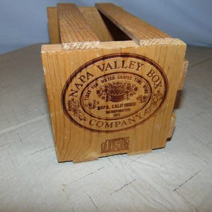 May include: A rectangular wooden crate with open slats and a natural light brown finish. The front panel features a circular dark brown brand stamp reading Napa Valley Box Company, Napa, California, Incorporated 1877, with a compact disc logo.