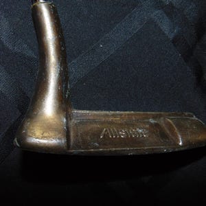 以下が含まれることがあります： 「Allstate」の文字が刻まれた、ヴィンテージのブロンズ製ゴルフパター。 パターは湾曲したネックと、平らな長方形のクラブヘッドを備えています。 パターは暗い背景に配置されています。