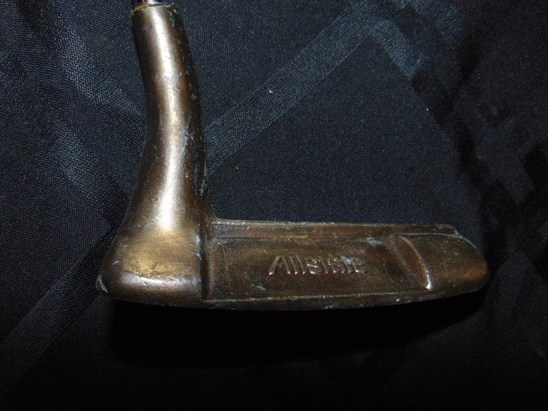 以下が含まれることがあります： 「Allstate」の文字が刻まれた、ヴィンテージのブロンズ製ゴルフパター。 パターは湾曲したネックと、平らな長方形のクラブヘッドを備えています。 パターは暗い背景に配置されています。