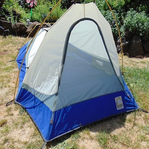 May include: A Sierra Designs dome tent featuring a blue base, light gray upper panels, and a large arched front entrance. The structure is supported by thin yellow poles, with a white mesh door panel visible inside the arched opening.