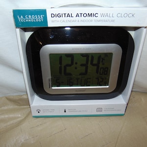 May include: A La Crosse Technology digital atomic wall clock in its original packaging. The clock features a black frame with a silver bezel, a digital display showing 12:34, the date 5-16, the day TUE, and an indoor temperature of 72 degrees.