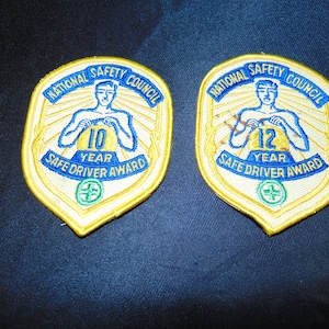 May include: Two yellow and blue National Safety Council Safe Driver Award patches. The left patch says "10 Year Safe Driver Award" and the right patch says "12 Year Safe Driver Award".