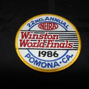 May include: A circular patch with a gold border, featuring the text "22nd Annual NHRA Winston World Finals 1986 Pomona, CA." in red, blue, and white. The patch is likely a collectible item or souvenir.