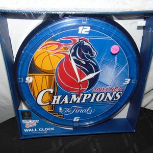 May include: A blue and red wall clock with a basketball theme. The clock features the 2004 NBA Champions logo, a basketball, and a trophy. The clock is in a clear plastic packaging with the words "Wall Clock" printed on the bottom.