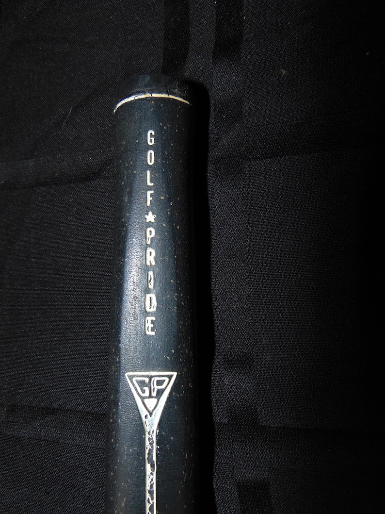 以下が含まれることがあります： 白い文字で「GOLF PRIDE」と書かれた黒いゴルフクラブグリップ。 グリップの上部には白いストライプがあり、三角形の中に「GP」が入った白いロゴが付いています。