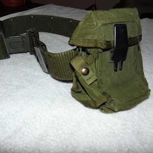 May include: An olive green military-style belt with a matching pouch. The belt features a metal buckle and a woven nylon strap. The pouch has a black plastic clip and a snap closure. The items are on a white surface.