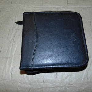 May include: A square black zippered case with a textured surface and a curved vertical seam detail on the left side. The case features a small embossed logo on the right side and a zipper pull tab visible at the top left corner.