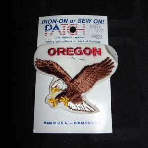 May include: An embroidered patch featuring a brown eagle with white and yellow accents, the word "OREGON" in red, and the text "IRON-ON or SEW ON!" on the packaging. Made in the U.S.A.