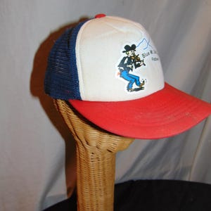 May include: A white, red, and blue trucker hat with a cartoon fiddler and the text "Blue Mt.  Oh. Fiddlers" on the front.