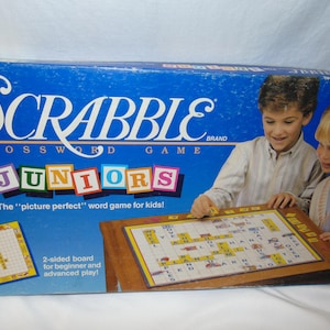 May include: A blue box with the words "SCRABBLE BRAND CROSSWORD GAME FOR JUNIORS" on the front. The box features a picture of two children playing the game. The game is described as "The "picture perfect" word game for kids!" and has a two-sided board for beginner and advanced play.