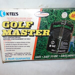 May include: A box for the K-Tel's Golf Master golf practice device. The box is red, white, and black with a picture of a golfer and the device. The device is black and has a dial with yards and meters. The box says "The Portable Driving Range That Improves Your Swing" and "Safe, Easy to Use, Saves Money".