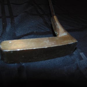 以下が含まれることがあります： ブロンズ色のヘッドと暗いシャフトが付いた、ヴィンテージのゴルフパター。パターヘッドは長方形です。クラブは使用感があり、摩耗や経年劣化の兆候が見られます。